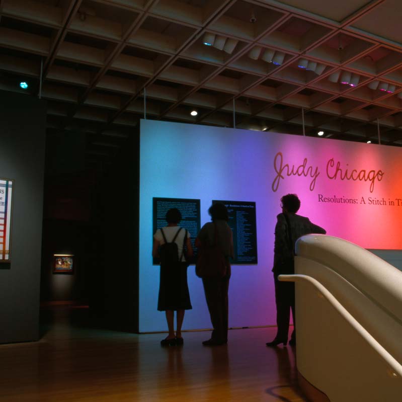 Color photograph of three people reading wall text in an exhibition with an artwork on the left and rainbow lights projected on the wall on the right