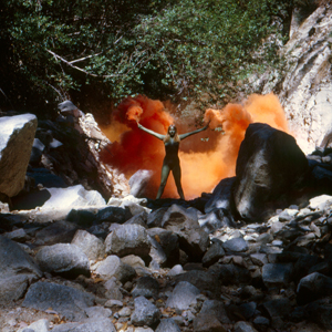 Color photograph of a woman standing on rocky terrain, surrounded by orange fireworks