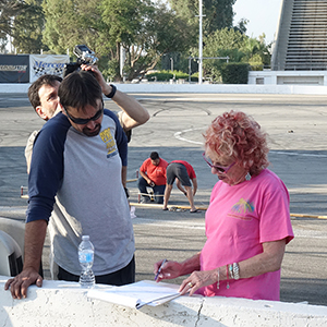Color photograph of Judy Chicago and Chris Souza standing and reviewing plans while Bill Bilowit documents the activities with a camera and Mary Costa and pyrotechnician Gabriel Garcia set up the smoke test on gray racetrack pavement in the background