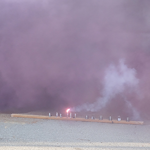 Color photograph of pink and purple smoke rising from a row of flares lined up on a wooden board