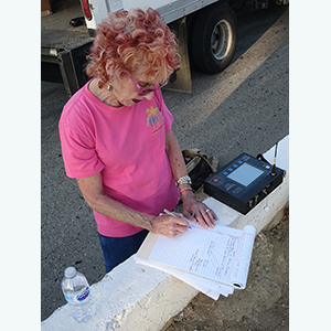 Color photograph of Judy Chicago standing at a white road divider making notes on a pad of paper next to a piece of equipment