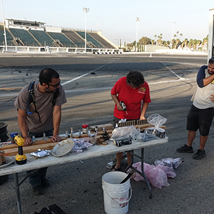 Color photograph of pyrotechnician Rusty Johnson, Mary Costa and Chris Souza preparing the smoke test at a folding table set up on the pavement of Orange Show Speedway with a truck nearby