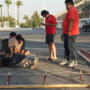 Color photograph of Rusty Johnson, Chris Souza, Mary Costa, and Gabriel Garcia connecting devices with electronic match to a computerized firing system on the pavement of Orange Show Speedway with grandstands in the background