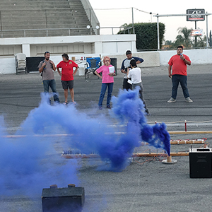 Color photograph of Rusty Johnson, Mary Costa, Judy Chicago, Chris Souza, and Gabriel Garcia standing and observing the blue smoke canister test on the pavement at Orange Show Speedway as Marilyn Loddi documents the activities with a camera