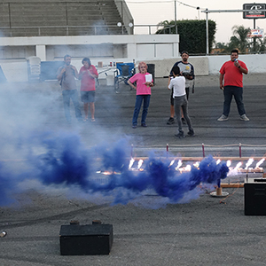 Color photograph of Rusty Johnson, Mary Costa, Judy Chicago, Chris Souza, and Gabriel Garcia standing and observing the blue smoke canister and white flares test on the pavement at Orange Show Speedway as Marilyn Loddi documents the activities with a camera