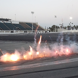 Color photograph of flares lighting in a row on the pavement at Orange Show Speedway with a grandstand in the background