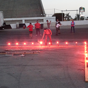 Color photograph of Rusty Johnson lighting a burned out flare in two perpendicular rows of orange flares as Mary Costa and Gabriel Garcia look on in the background and Marilyn Loddi and Bill Bilowit document Chris Souza and Judy Chicago with cameras on the pavement at Orange Show Speedway