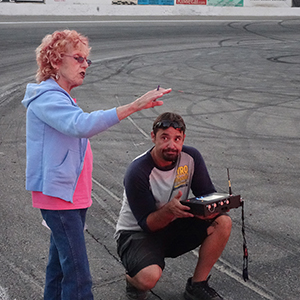 Color photograph of Judy Chicago standing and gesturing while Chris Souza is kneeling and holding a piece of electronic equipment on the pavement at Orange Show Speedway