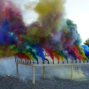 Color photograph of rainbow-colored smoke rising from canisters mounted on a rectangular wooden structure in a gravel lot