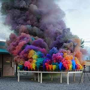 Color photograph of rainbow-colored smoke rising from canisters mounted on a rectangular wooden structure in a gravel lot with buildings in the background and a tripod in the foreground