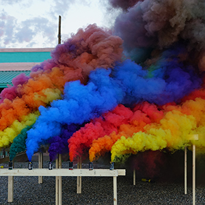 Color photograph of rainbow-colored smoke rising from canisters mounted on a rectangular wooden structure in a gravel lot with buildings in the background