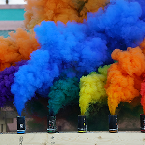 Color photograph of rainbow-colored smoke rising from a row of canisters mounted on a rectangular wooden structure