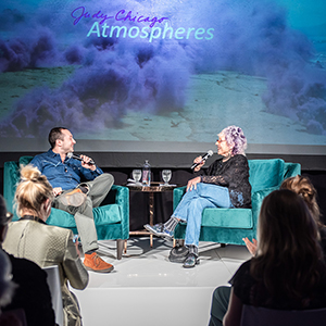 Color photograph of Judy Chicago and Alex Gartenfeld sitting in teal chairs on a dais in front of an audience with a slide projection of purple smoke behind them