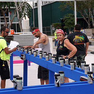 Color photograph of Rusty Johnson, Chris Souza, and Mary Costa installing smoke and fireworks canisters on a blue structure in a plaza with buildings in the background