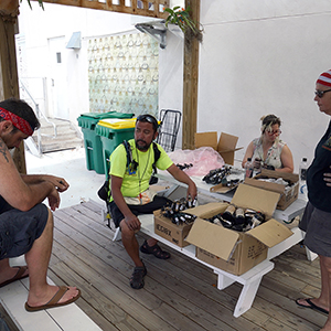 Color photograph of Chris Souza, Gabriel Garcia, Rusty Johnson, an unknown woman, and Mary Costa sitting on white benches with boxes of smoke canisters and fireworks
