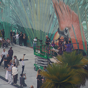Color photograph of Judy Chicago delivering remarks from a scaffold with Donald Woodman, Mary Costa, Chris Souza, Vanessa Olvera, and Gabriel Garcia and others in front of a large mural