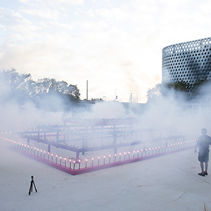 Color photograph of white smoke filling a plaza above a rectangular structure lined with flares with Rusty Johnson lighting the bottom tier of smoke flares