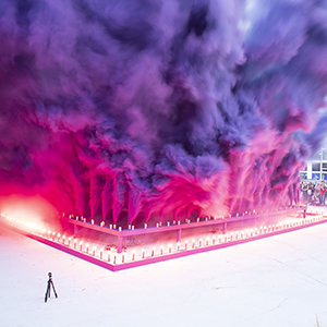 Color photograph of purple and pink smoke emerging from flares on a multicolored rectangular structure in a plaza as a crowd of people looks on