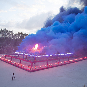 Color photograph of blue smoke emerging from flares on a multi colored rectangular structure in a plaza as a crowd of people looks on