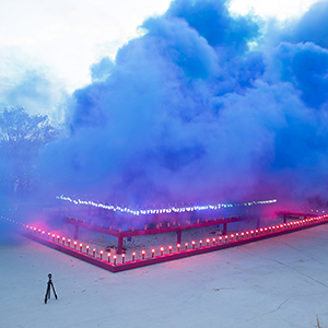 Color photograph of blue smoke emerging from flares on a multi colored rectangular structure in a plaza