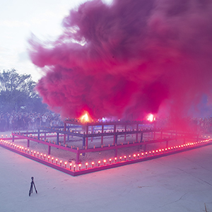 Color photograph of pink smoke emerging from flares on a multicolored rectangular structure in a plaza as a crowd of people looks on