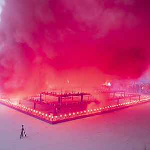Color photograph of pink smoke emerging from flares on a multicolored rectangular structure in a plaza as a crowd of people looks on