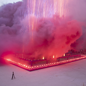 Color photograph of pink and purple smoke emerging from flares on a multicolored rectangular structure with fireworks in a plaza as a crowd of people looks on