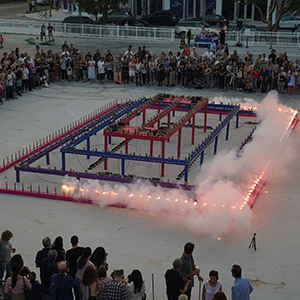 Color photograph of white smoke rising from one side of a multicolored, rectangular structure in a plaza as Rusty Johnson lights the bottom tier of smoke flares and a crowd of people looks on