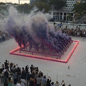 Color photograph of purple smoke emerging from flares on a multi colored rectangular structure in a plaza as a crowd of people looks on