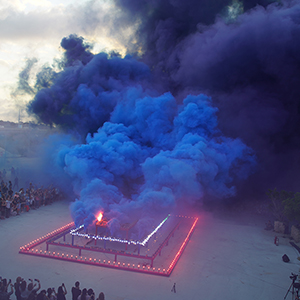 Color photograph of blue and purple smoke emerging from flares on a multicolored rectangular structure in a plaza as a crowd of people looks on