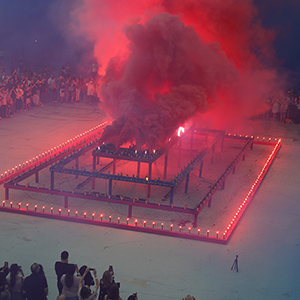 Color photograph of red smoke emerging from flares on a multicolored rectangular structure in a plaza as a crowd of people looks on