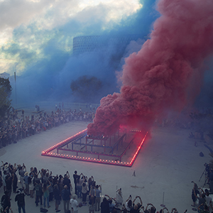 Color photograph of red and blue smoke emerging from flares on a multicolored rectangular structure in a plaza as a crowd of people looks on