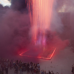 Color photograph of pink smoke and fireworks emerging from flares on a multicolored rectangular structure in a plaza as a crowd of people looks on