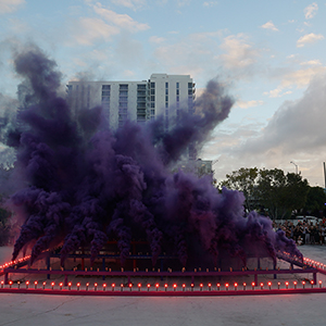 Color photograph of purple smoke emerging from flares on a multicolored rectangular structure in a plaza as a crowd of people looks on