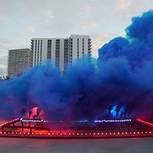 Color photograph of blue smoke emerging from flares on a multicolored rectangular structure in a plaza as a crowd of people looks on