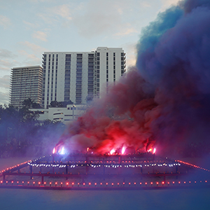 Color photograph of red and blue smoke emerging from flares on a multicolored rectangular structure in a plaza as a crowd of people looks on