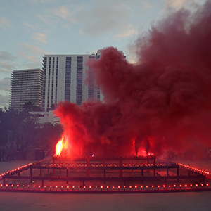 Color photograph of red smoke emerging from flares on a multicolored rectangular structure in a plaza as a crowd of people looks on
