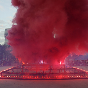 Color photograph of red smoke emerging from flares on a multi colored rectangular structure in a plaza as a crowd of people looks on