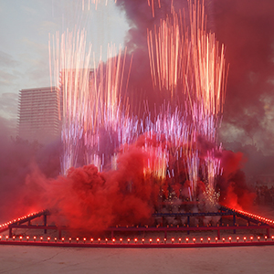 Color photograph of red smoke and fireworks emerging from flares on a multicolored rectangular structure in a plaza as a crowd of people looks on