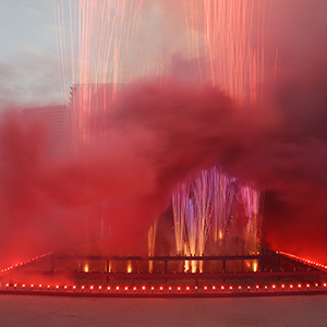 Color photograph of red and purple smoke and fireworks emerging from flares on a multicolored rectangular structure in a plaza as a crowd of people looks on
