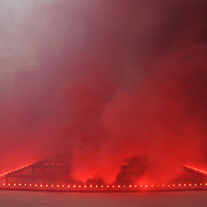Color photograph of red and purple smoke emerging from flares on a multicolored rectangular structure in a plaza as a crowd of people looks on