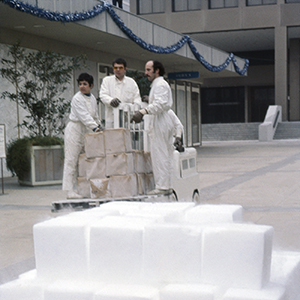 Color photograph of Judy Chicago, Eric Orr, and Lloyd Hamrol riding on a forklift loaded with blocks wrapped in paper behind a stack of white blocks