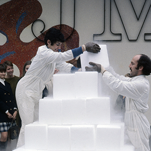 Color photograph of Judy Chicago and Lloyd Hamrol stacking white blocks in a pyramid shape as other people look on