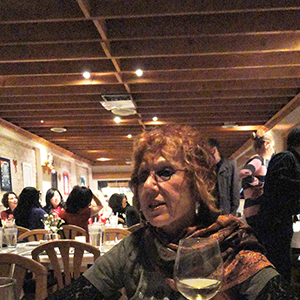 Color photograph of Judy Chicago sitting at a table with a glass of wine and a pyramid of white sugar cubes with other people in the background