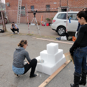 Color photograph of a person kneeling next to a small pyramid of white blocks as a group of people look on