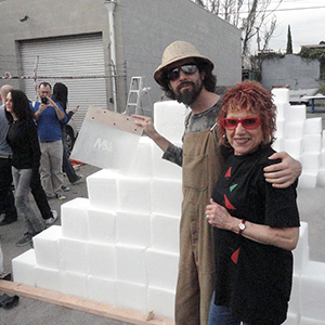 Color photograph of Oliver Hess and Judy Chicago standing in front of two pyramids of white blocks with other people standing nearby