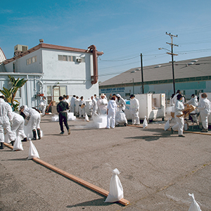 Color photograph of a group of people dressed in white installing white blocks in a paved lot lined with wooden beams held down with sandbags