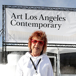 Color photograph of Judy Chicago, dressed in white, standing in front of a large banner