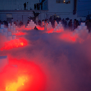 Color photograph of two rows of pyramids of white blocks with fog and red lights emanating from them and people standing around in the evening