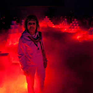 Color photograph of Judy Chicago standing in front of pyramids of blocks glowing red and emanating fog